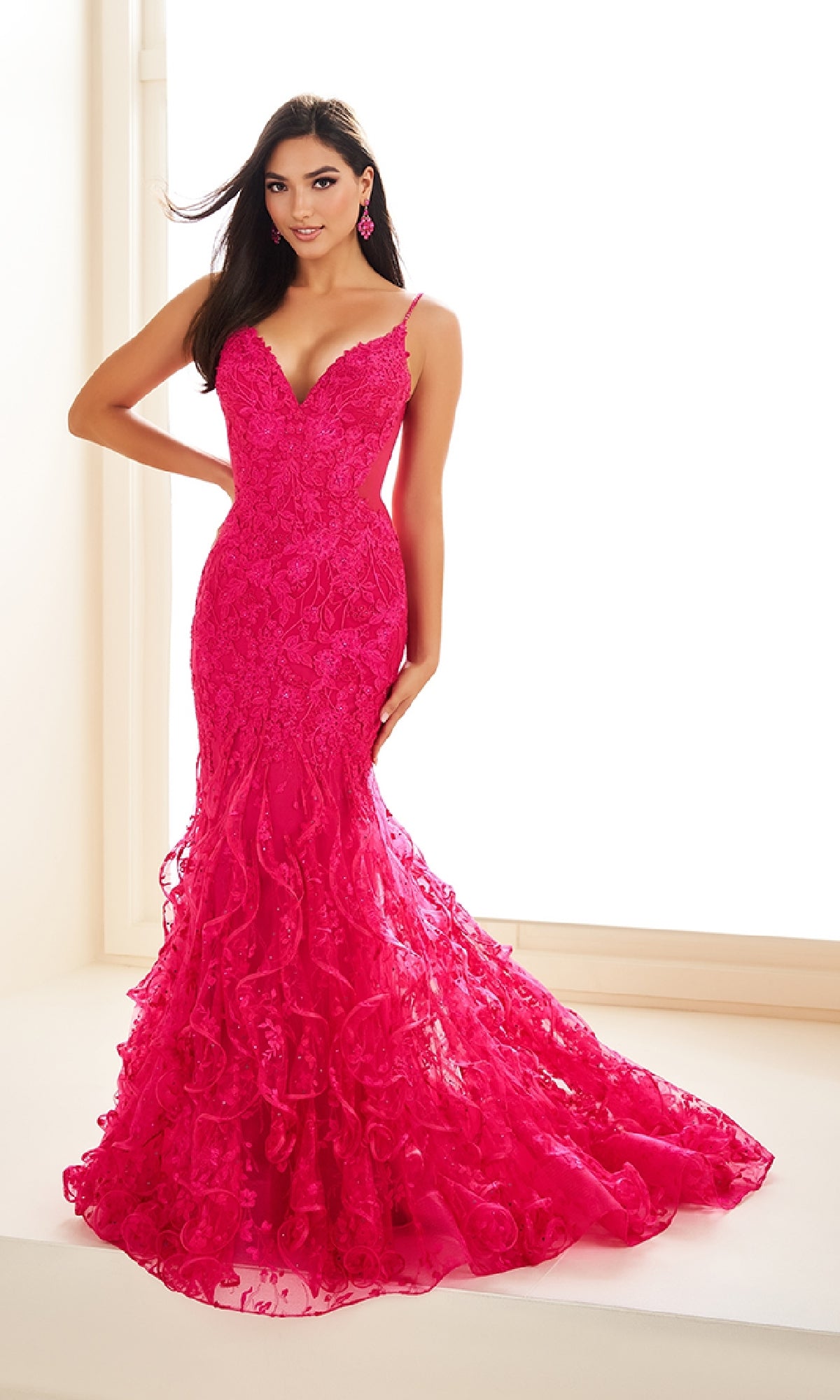 Woman wearing the Ellie Wilde EW36041 long prom dress, a bright pink, form-fitting lace mermaid gown with a v-neckline and thin straps, standing indoors against a white background.